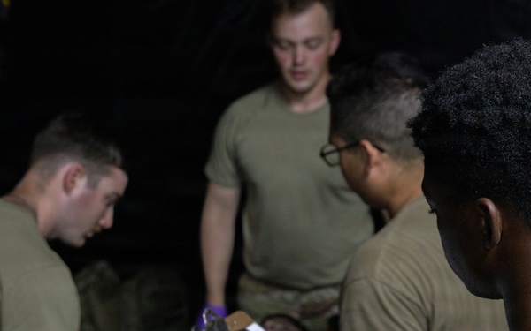 The image shows a group of military personnel in olive drab t-shirts engaged in what appears to be a medical training exercise. One individual is wearing purple medical gloves and appears to be performing a procedure or assessment on a person lying down, while another holds a clipboard with a checklist. The scene is set in a low-light environment consistent with a field training scenario.