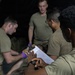 The image shows a group of military personnel in olive drab t-shirts engaged in what appears to be a medical training exercise. One individual is wearing purple medical gloves and appears to be performing a procedure or assessment on a person lying down, while another holds a clipboard with a checklist. The scene is set in a low-light environment consistent with a field training scenario.