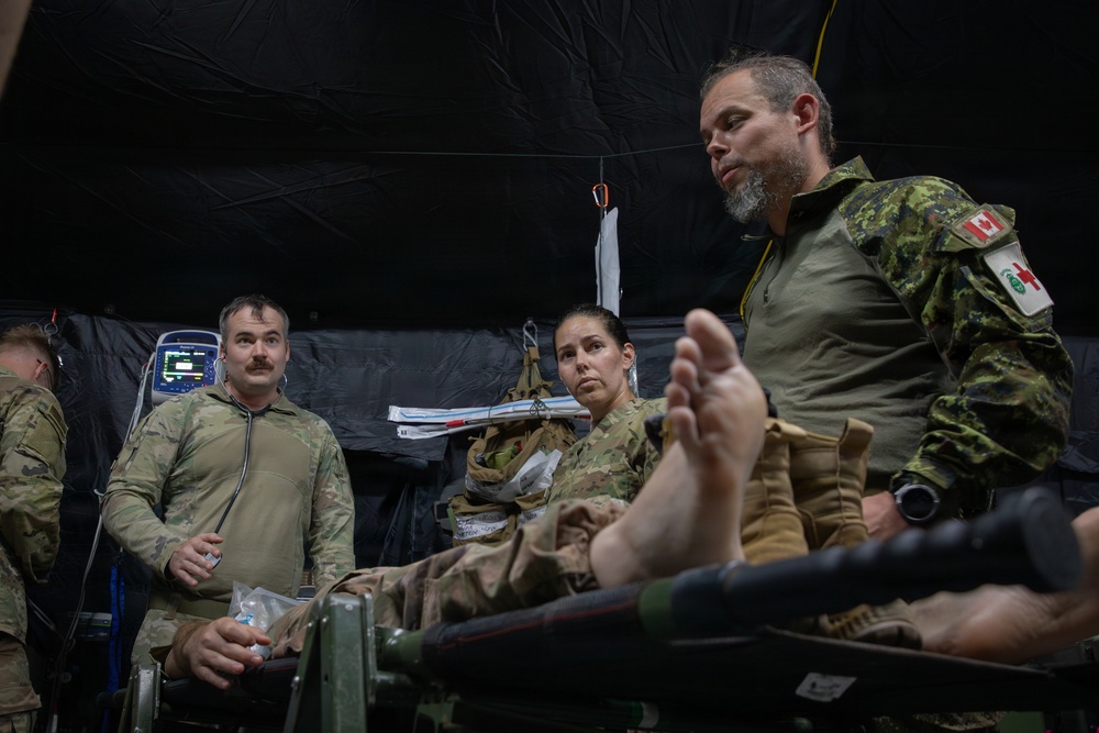 Military medical personnel are gathered inside a field hospital tent, attending to a patient on a stretcher. At least three uniformed individuals are visible, including one wearing a Canadian Armed Forces uniform with a visible Canadian flag patch. The setting appears to be a tactical field medical exercise or real-world medical operation.