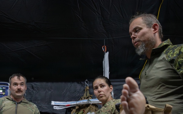 Military medical personnel are gathered inside a field hospital tent, attending to a patient on a stretcher. At least three uniformed individuals are visible, including one wearing a Canadian Armed Forces uniform with a visible Canadian flag patch. The setting appears to be a tactical field medical exercise or real-world medical operation.