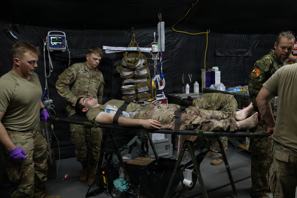 A military medical treatment scenario taking place inside a field hospital tent, where personnel in military uniforms are attending to a patient lying on a stretcher. Multiple medical providers, including at least one individual in a different camouflage pattern (possibly Canadian Armed Forces), are working around the patient. Medical equipment including a vital signs monitor, IV drip, and various supplies are visible.
