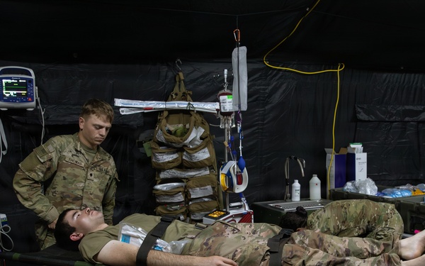 A military medical treatment scenario taking place inside a field hospital tent, where personnel in military uniforms are attending to a patient lying on a stretcher. Multiple medical providers, including at least one individual in a different camouflage pattern (possibly Canadian Armed Forces), are working around the patient. Medical equipment including a vital signs monitor, IV drip, and various supplies are visible.
