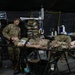 A military medical treatment scenario taking place inside a field hospital tent, where personnel in military uniforms are attending to a patient lying on a stretcher. Multiple medical providers, including at least one individual in a different camouflage pattern (possibly Canadian Armed Forces), are working around the patient. Medical equipment including a vital signs monitor, IV drip, and various supplies are visible.