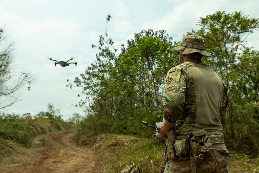 A military personnel in full camouflage uniform and boonie hat is operating a small quadcopter drone in a rural, vegetated outdoor environment. The operator is holding a remote controller and observing the drone in flight. The setting appears to be a training or reconnaissance operation in a tropical or subtropical landscape.