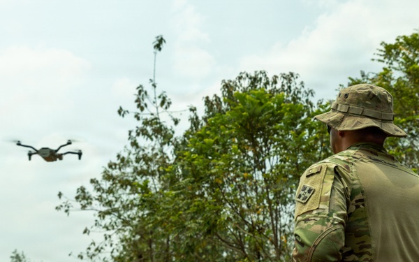 A military personnel in full camouflage uniform and boonie hat is operating a small quadcopter drone in a rural, vegetated outdoor environment. The operator is holding a remote controller and observing the drone in flight. The setting appears to be a training or reconnaissance operation in a tropical or subtropical landscape.