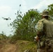 A military personnel in full camouflage uniform and boonie hat is operating a small quadcopter drone in a rural, vegetated outdoor environment. The operator is holding a remote controller and observing the drone in flight. The setting appears to be a training or reconnaissance operation in a tropical or subtropical landscape.
