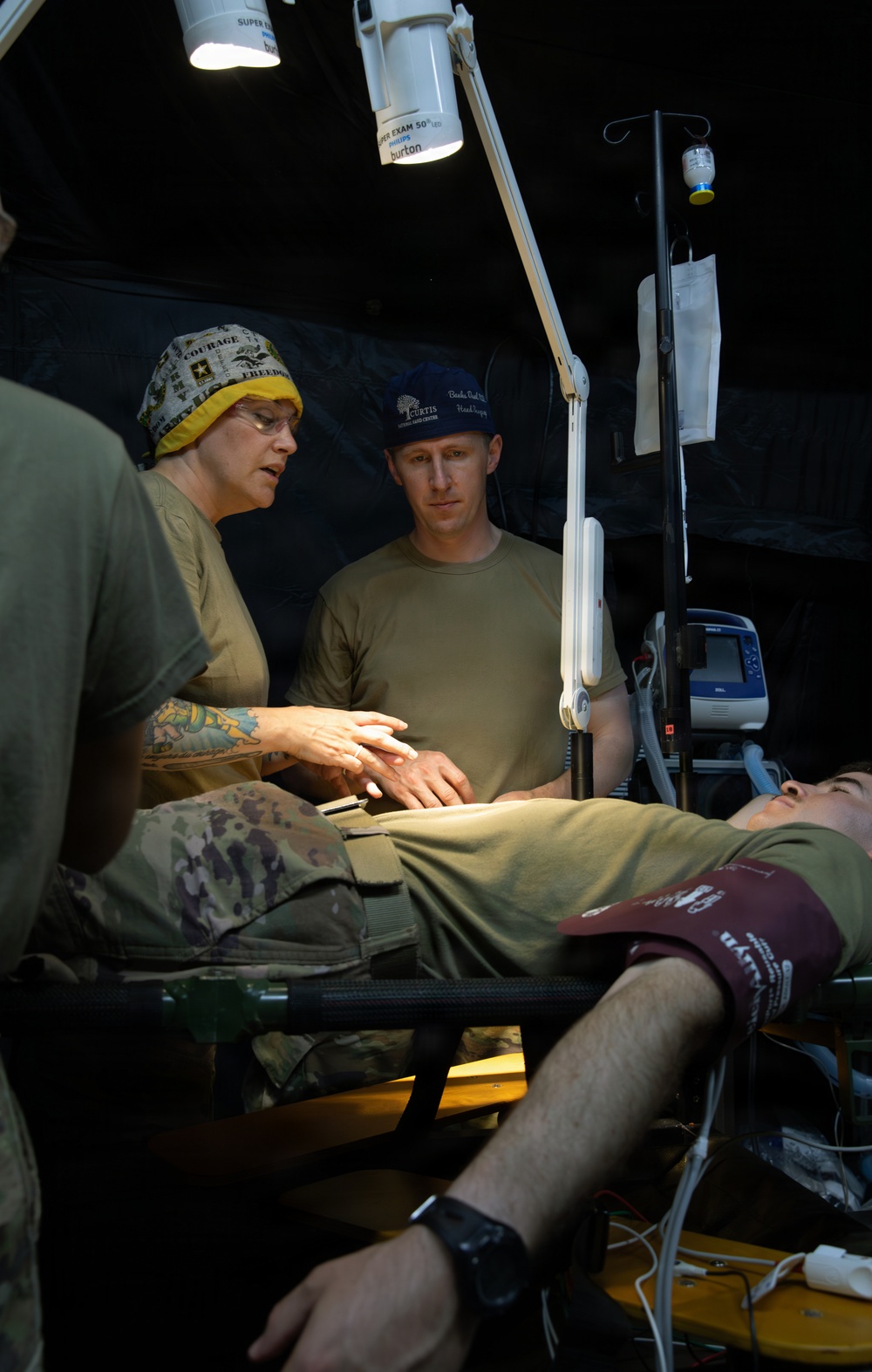 A field medical treatment scene showing military personnel providing care to a patient lying on a stretcher inside what appears to be a military medical tent. Two medical providers are attending to the patient, with one wearing a surgical cap bearing 'COURAGE' and 'FREEDOM' text and the other wearing a blue cap. Medical equipment including an IV stand, monitoring devices, and examination lights are visible.