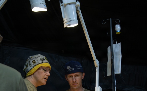 A field medical treatment scene showing military personnel providing care to a patient lying on a stretcher inside what appears to be a military medical tent. Two medical providers are attending to the patient, with one wearing a surgical cap bearing 'COURAGE' and 'FREEDOM' text and the other wearing a blue cap. Medical equipment including an IV stand, monitoring devices, and examination lights are visible.