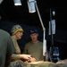 A field medical treatment scene showing military personnel providing care to a patient lying on a stretcher inside what appears to be a military medical tent. Two medical providers are attending to the patient, with one wearing a surgical cap bearing 'COURAGE' and 'FREEDOM' text and the other wearing a blue cap. Medical equipment including an IV stand, monitoring devices, and examination lights are visible.