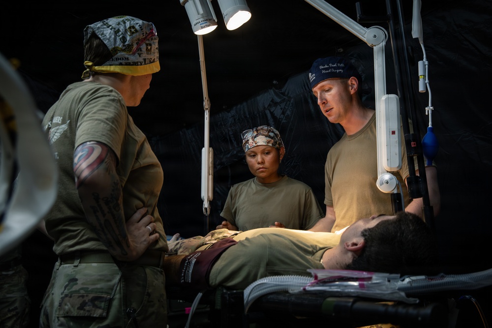 Military medical personnel are attending to a patient lying on a stretcher or medical table in what appears to be a field medical tent or forward surgical unit. Three individuals in military attire and surgical caps are gathered around the patient under a portable surgical lamp. The setting suggests a combat support hospital or tactical field surgery environment.