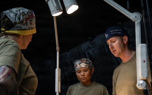 Military medical personnel are attending to a patient lying on a stretcher or medical table in what appears to be a field medical tent or forward surgical unit. Three individuals in military attire and surgical caps are gathered around the patient under a portable surgical lamp. The setting suggests a combat support hospital or tactical field surgery environment.