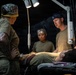 Military medical personnel are attending to a patient lying on a stretcher or medical table in what appears to be a field medical tent or forward surgical unit. Three individuals in military attire and surgical caps are gathered around the patient under a portable surgical lamp. The setting suggests a combat support hospital or tactical field surgery environment.
