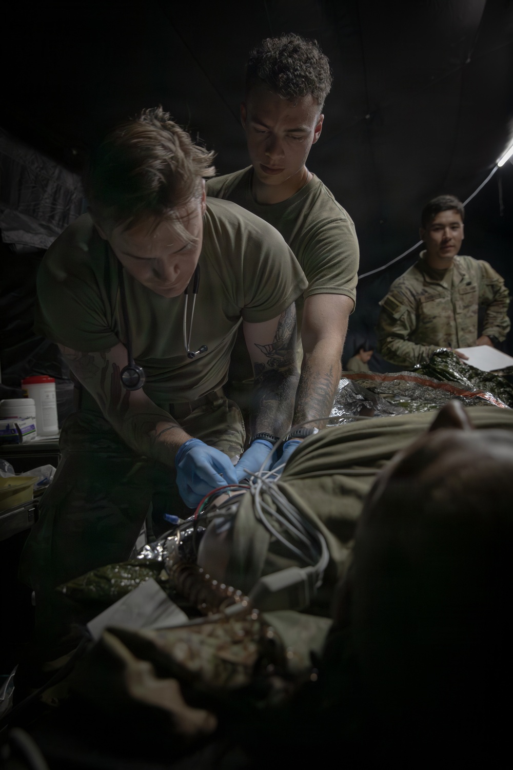 Two military personnel in olive drab t-shirts are performing what appears to be a medical training exercise or treatment on a patient lying on a surface covered with a thermal/mylar blanket. One individual is wearing blue medical gloves and a stethoscope, suggesting a medic or corpsman role. A third uniformed service member in full camouflage uniform observes from the background, possibly in a supervisory or evaluator capacity.