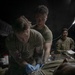 Two military personnel in olive drab t-shirts are performing what appears to be a medical training exercise or treatment on a patient lying on a surface covered with a thermal/mylar blanket. One individual is wearing blue medical gloves and a stethoscope, suggesting a medic or corpsman role. A third uniformed service member in full camouflage uniform observes from the background, possibly in a supervisory or evaluator capacity.