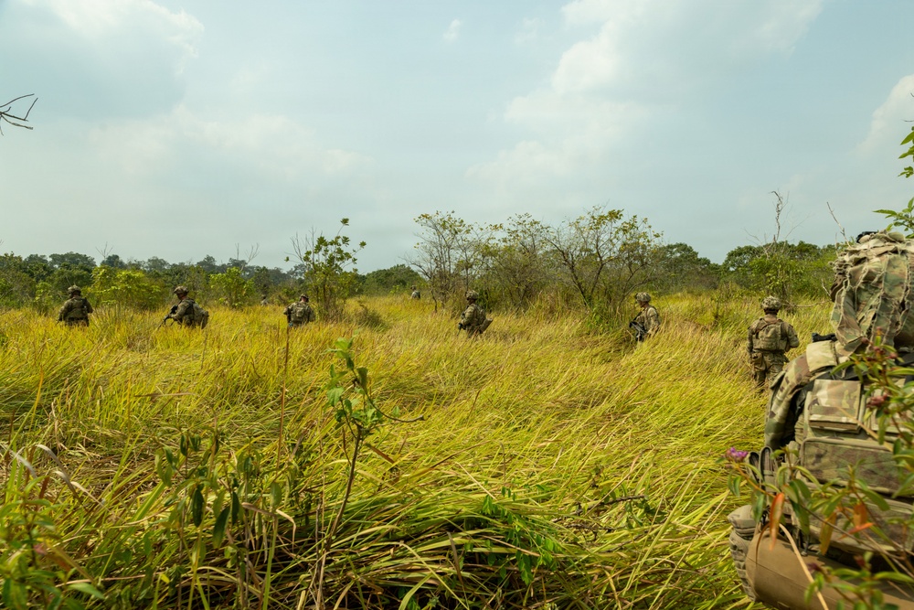 A group of military personnel in camouflage uniforms are conducting a patrol or field exercise through tall grassland terrain with scattered trees and shrubs. The soldiers are spread out in a tactical formation moving away from the camera's perspective. The environment appears to be a tropical or subtropical savanna-like landscape under a partly cloudy sky.