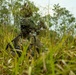 A heavily camouflaged soldier is positioned low in tall grass, wearing full tactical gear including a helmet with foliage attached and face paint. The soldier appears to be in a concealed observation or overwatch position in a lush, vegetated environment. The image captures a military field exercise or operational scenario emphasizing stealth and concealment techniques.