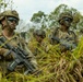 Three military personnel in full combat gear are positioned in tall grass, likely conducting a field training exercise or tactical operation. All three wear camouflage face paint, tactical helmets, body armor, and are equipped with assault rifles. The setting appears to be a tropical or subtropical environment based on the vegetation.