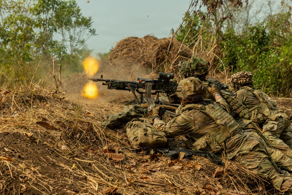 Military personnel in camouflage uniforms are in a prone firing position, operating a belt-fed machine gun that is actively firing, producing a visible muzzle flash and flame. The soldiers appear to be conducting a live-fire training exercise or combat operation in a dry, scrubland environment. At least two to three personnel are visible, with one operating the weapon and others in support positions.