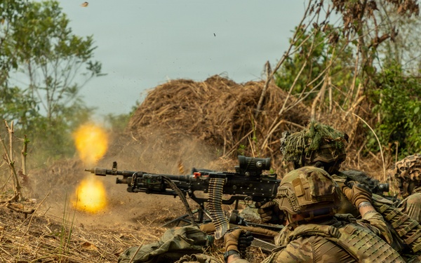 Military personnel in camouflage uniforms are in a prone firing position, operating a belt-fed machine gun that is actively firing, producing a visible muzzle flash and flame. The soldiers appear to be conducting a live-fire training exercise or combat operation in a dry, scrubland environment. At least two to three personnel are visible, with one operating the weapon and others in support positions.