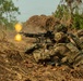 Military personnel in camouflage uniforms are in a prone firing position, operating a belt-fed machine gun that is actively firing, producing a visible muzzle flash and flame. The soldiers appear to be conducting a live-fire training exercise or combat operation in a dry, scrubland environment. At least two to three personnel are visible, with one operating the weapon and others in support positions.