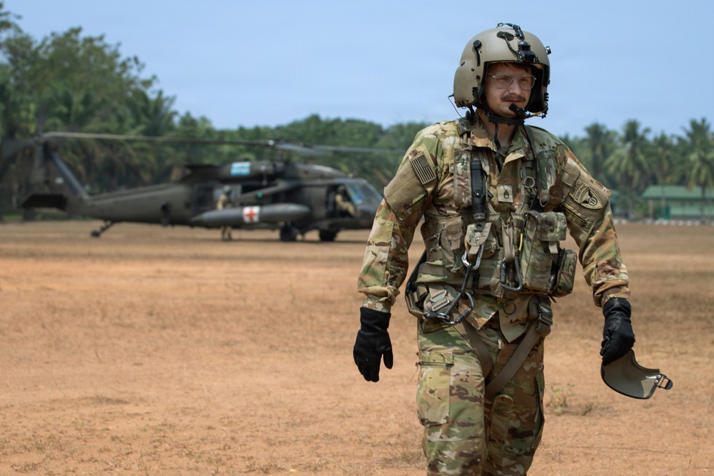 A U.S. Army soldier in full flight crew gear walks across a dirt landing zone, with a UH-60 Black Hawk medevac helicopter visible in the background. The soldier is wearing OCP (Operational Camouflage Pattern) uniform, a flight helmet with communications equipment, tactical vest, and black flight gloves. The tropical vegetation in the background suggests a deployment in a tropical or subtropical region.