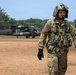 A U.S. Army soldier in full flight crew gear walks across a dirt landing zone, with a UH-60 Black Hawk medevac helicopter visible in the background. The soldier is wearing OCP (Operational Camouflage Pattern) uniform, a flight helmet with communications equipment, tactical vest, and black flight gloves. The tropical vegetation in the background suggests a deployment in a tropical or subtropical region.
