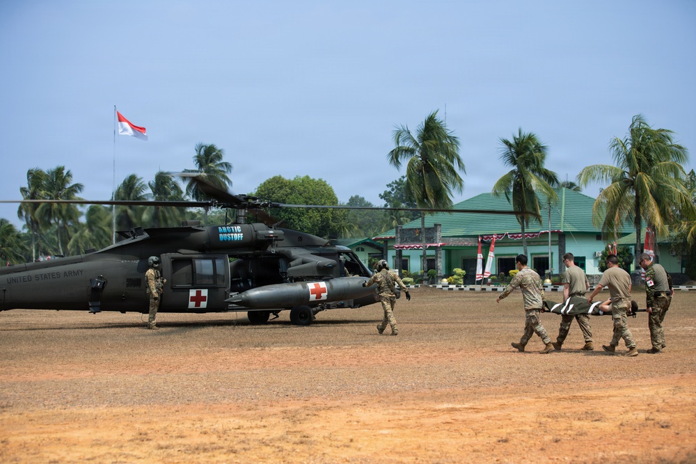 A military medical evacuation (MEDEVAC) exercise is taking place on a dirt landing zone, involving United States Army personnel and what appears to be a joint operation with Indonesian forces, indicated by the Indonesian flag visible in the background. Two UH-60 Black Hawk helicopters marked with Red Cross symbols and labeled 'Arctic Dustoff' and 'United States Army' are on the ground. Personnel are carrying a litter (stretcher) with a simulated casualty toward one of the helicopters.