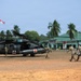 A military medical evacuation (MEDEVAC) exercise is taking place on a dirt landing zone, involving United States Army personnel and what appears to be a joint operation with Indonesian forces, indicated by the Indonesian flag visible in the background. Two UH-60 Black Hawk helicopters marked with Red Cross symbols and labeled 'Arctic Dustoff' and 'United States Army' are on the ground. Personnel are carrying a litter (stretcher) with a simulated casualty toward one of the helicopters.