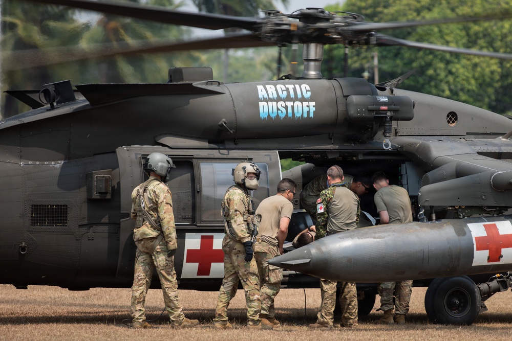 A military medical evacuation (MEDEVAC) helicopter marked 'Arctic Dustoff' is shown on the ground with personnel gathered around it. The UH-60 Black Hawk helicopter displays Red Cross markings, indicating its medical role. Several soldiers in various uniforms and flight gear appear to be loading or attending to a patient or equipment through the side door.