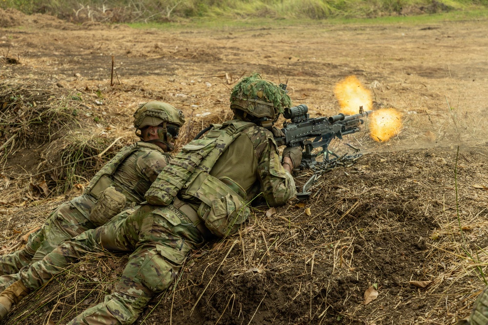 Two soldiers in full combat gear are positioned in a prone firing position in a shallow earthen trench or berm. The forward soldier is actively firing a machine gun, with a visible muzzle flash and ejected debris captured at the moment of discharge. The setting appears to be a training range or field exercise environment.