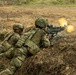 Two soldiers in full combat gear are positioned in a prone firing position in a shallow earthen trench or berm. The forward soldier is actively firing a machine gun, with a visible muzzle flash and ejected debris captured at the moment of discharge. The setting appears to be a training range or field exercise environment.