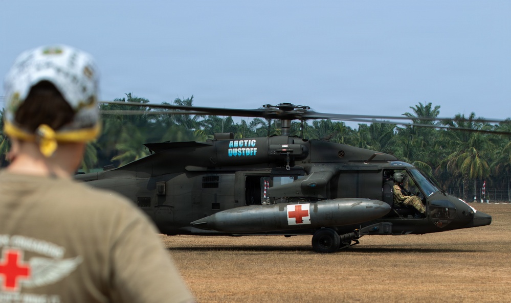 A military medical evacuation (MEDEVAC) UH-60 Black Hawk helicopter marked 'Arctic Dustoff' is on the ground at what appears to be a tropical location. A soldier wearing a red cross-marked shirt and helmet is observing the helicopter from the foreground. The aircraft bears red cross markings indicating its medical/MEDEVAC role.