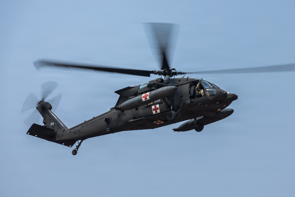 A United States Army UH-60 Black Hawk helicopter configured as a medevac (medical evacuation) aircraft is captured in flight against an overcast blue-gray sky. The helicopter displays prominent red cross markings on its fuselage, identifying it as a medical transport. The rotor blades show motion blur, indicating active flight.