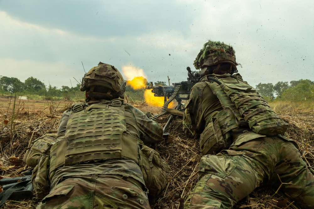 Two soldiers in full combat gear are in a prone firing position in an open field, operating what appears to be a belt-fed machine gun with a visible muzzle flash. Both personnel are wearing modern camouflage uniforms and tactical vests, suggesting a military training exercise or live-fire drill. The setting is a grassy, open terrain with trees visible in the background under an overcast sky.