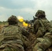 Two soldiers in full combat gear are in a prone firing position in an open field, operating what appears to be a belt-fed machine gun with a visible muzzle flash. Both personnel are wearing modern camouflage uniforms and tactical vests, suggesting a military training exercise or live-fire drill. The setting is a grassy, open terrain with trees visible in the background under an overcast sky.
