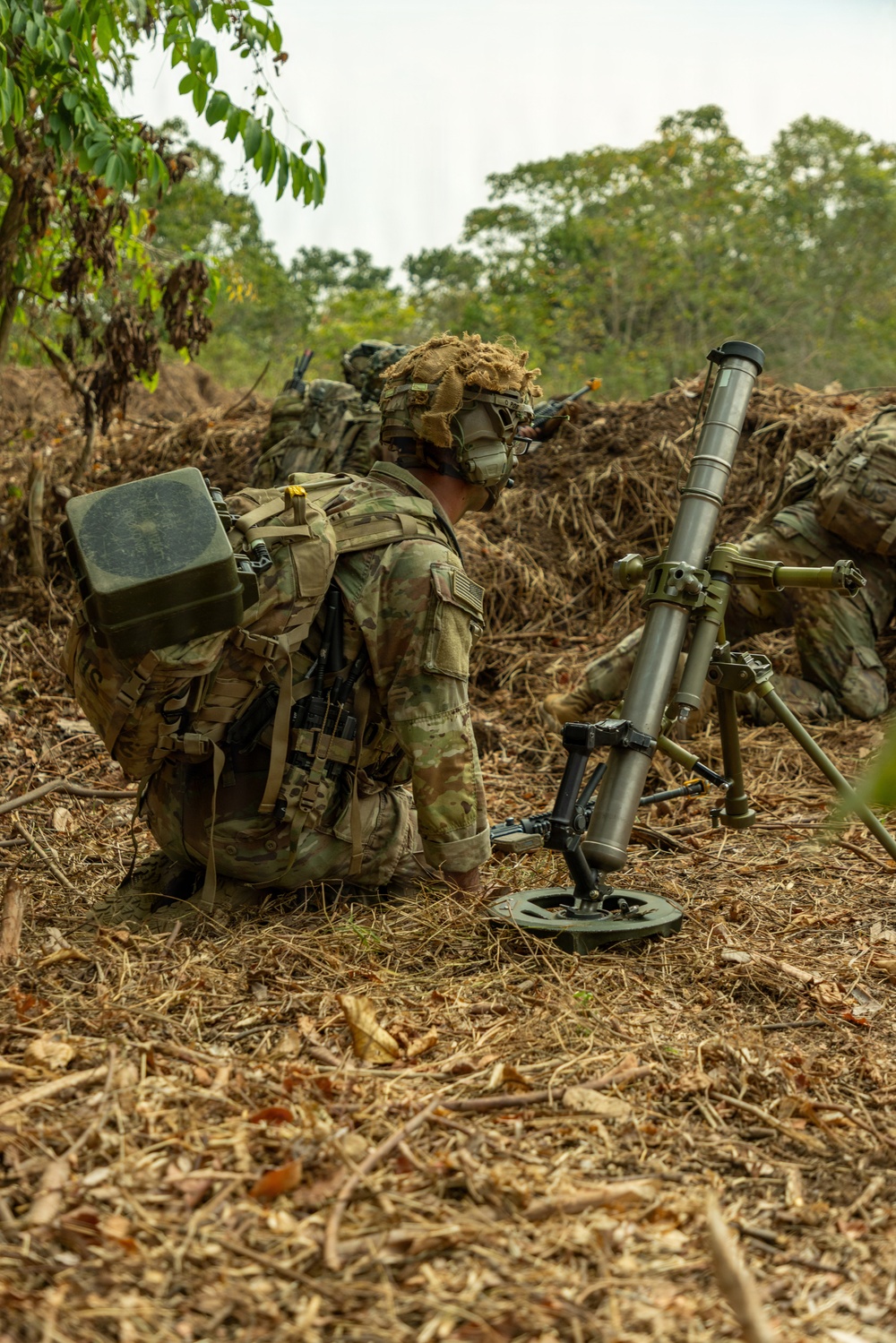 The image shows military personnel in full combat gear positioned in a field environment with dense vegetation in the background. A mortar system, identifiable by its tube, bipod, and baseplate, is set up beside the primary soldier who is kneeling and appears to be observing or communicating. The soldiers are wearing MultiCam (OCP) camouflage uniforms with helmets adorned with burlap/ghillie material, consistent with U.S. Army equipment and dress.