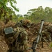 The image shows military personnel in full combat gear positioned in a field environment with dense vegetation in the background. A mortar system, identifiable by its tube, bipod, and baseplate, is set up beside the primary soldier who is kneeling and appears to be observing or communicating. The soldiers are wearing MultiCam (OCP) camouflage uniforms with helmets adorned with burlap/ghillie material, consistent with U.S. Army equipment and dress.
