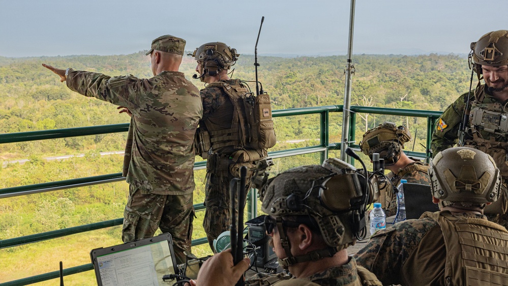 A group of military personnel in full combat gear are positioned on an elevated observation platform overlooking a dense jungle or forested area. One soldier in OCP/multicam uniform is pointing toward the treeline, apparently directing attention to a specific area, while others monitor communications equipment and laptops. The scene appears to be a coordinated military exercise or operation command post.