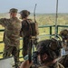 A group of military personnel in full combat gear are positioned on an elevated observation platform overlooking a dense jungle or forested area. One soldier in OCP/multicam uniform is pointing toward the treeline, apparently directing attention to a specific area, while others monitor communications equipment and laptops. The scene appears to be a coordinated military exercise or operation command post.
