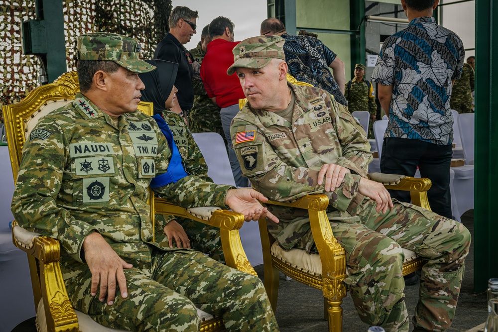 Two military officers — one from the Indonesian Army (TNI AD) and one from the U.S. Army — are seated in ornate gold chairs engaged in conversation, likely during a bilateral military event or exercise. The Indonesian officer wears the name tape 'NAUDI' with two-star rank insignia, while the U.S. officer displays Airborne and 101st Airborne Division patches along with a brigadier general star. Several other personnel and civilians are visible in the background.