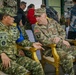 Two military officers — one from the Indonesian Army (TNI AD) and one from the U.S. Army — are seated in ornate gold chairs engaged in conversation, likely during a bilateral military event or exercise. The Indonesian officer wears the name tape 'NAUDI' with two-star rank insignia, while the U.S. officer displays Airborne and 101st Airborne Division patches along with a brigadier general star. Several other personnel and civilians are visible in the background.