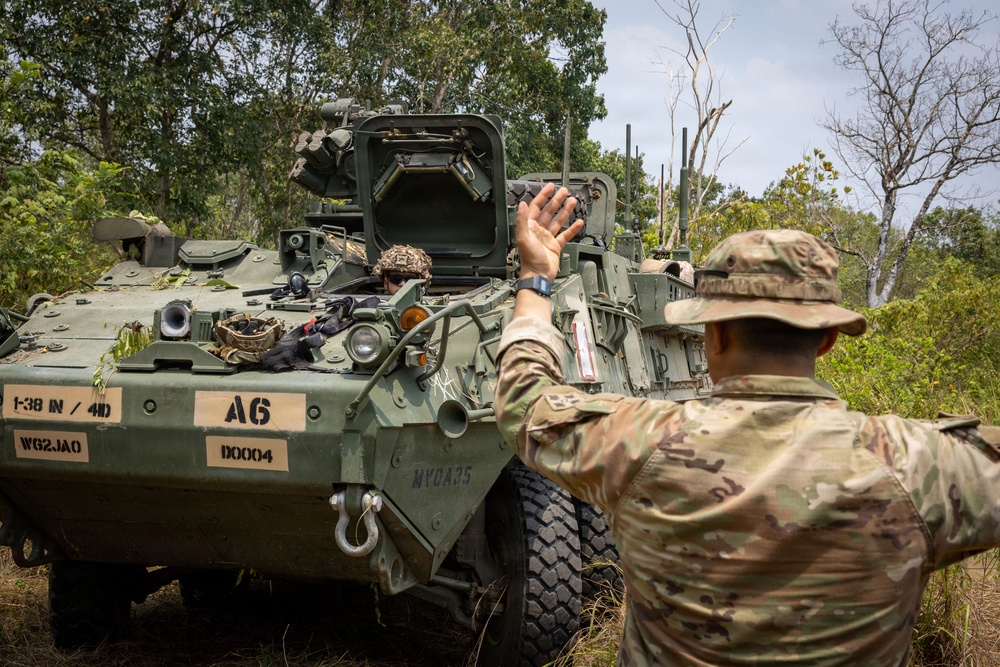 A soldier in MultiCam/OCP camouflage uniform is directing or signaling to crew members inside a Stryker armored fighting vehicle marked 'A6' belonging to 1-38 Infantry, 4th Division. The scene appears to be a field training exercise or operational movement in a wooded, tropical or subtropical environment. The vehicle displays unit markings including '1-38 IN / 4D', 'WG2JAO', and 'D0004'.