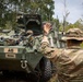 A soldier in MultiCam/OCP camouflage uniform is directing or signaling to crew members inside a Stryker armored fighting vehicle marked 'A6' belonging to 1-38 Infantry, 4th Division. The scene appears to be a field training exercise or operational movement in a wooded, tropical or subtropical environment. The vehicle displays unit markings including '1-38 IN / 4D', 'WG2JAO', and 'D0004'.