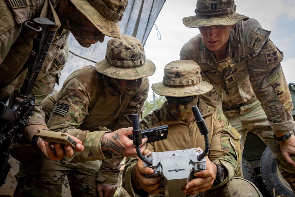 Four U.S. Army soldiers in multicam uniforms and boonie hats are gathered around a small drone or UAS (Unmanned Aerial System) controller/device. The soldiers appear to be conducting training or a briefing on drone operation, with one soldier holding the device while others observe and interact with it. Visible rank insignia and unit patches indicate this is an organized military activity.
