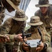 Four U.S. Army soldiers in multicam uniforms and boonie hats are gathered around a small drone or UAS (Unmanned Aerial System) controller/device. The soldiers appear to be conducting training or a briefing on drone operation, with one soldier holding the device while others observe and interact with it. Visible rank insignia and unit patches indicate this is an organized military activity.
