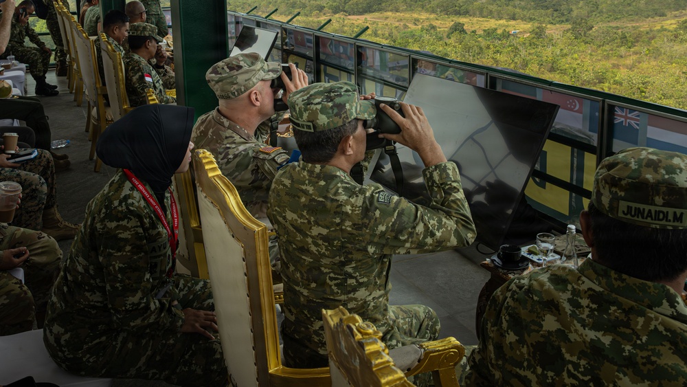 Military personnel from multiple nations are gathered in an elevated observation platform, using binoculars to observe what appears to be a joint military exercise or demonstration. The setting includes a viewing area overlooking a green, hilly landscape, with multiple national flags visible on display screens or panels. The presence of diverse camouflage uniforms and insignia suggests a multinational military event.