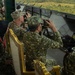 Military personnel from multiple nations are gathered in an elevated observation platform, using binoculars to observe what appears to be a joint military exercise or demonstration. The setting includes a viewing area overlooking a green, hilly landscape, with multiple national flags visible on display screens or panels. The presence of diverse camouflage uniforms and insignia suggests a multinational military event.