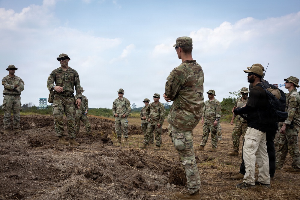 A group of military personnel in camouflage uniforms are gathered in a semi-circle on open, disturbed terrain, appearing to receive a briefing or after-action review. One soldier facing away from the camera appears to be addressing the group, while another in a wide-brim hat stands opposite. One civilian or contractor in tan pants and a black vest with a radio is also present among the group.