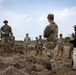 A group of military personnel in camouflage uniforms are gathered in a semi-circle on open, disturbed terrain, appearing to receive a briefing or after-action review. One soldier facing away from the camera appears to be addressing the group, while another in a wide-brim hat stands opposite. One civilian or contractor in tan pants and a black vest with a radio is also present among the group.