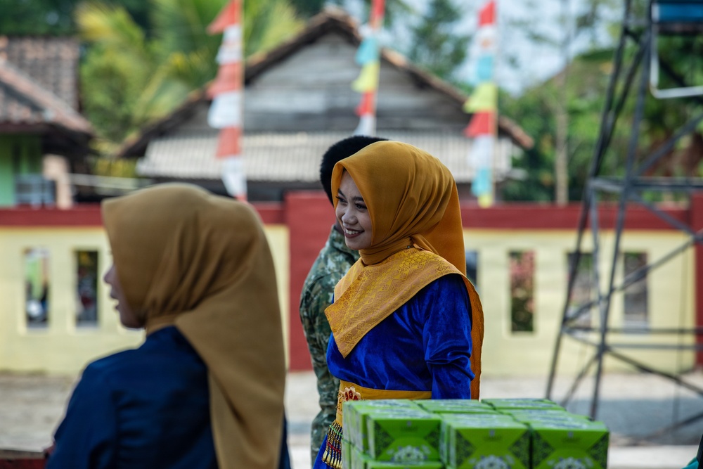 The image shows two women wearing hijabs and traditional-style clothing in an outdoor setting, with one woman smiling and holding green boxes. The background features colorful flags/bunting and buildings with red and yellow painted walls, suggesting a community or celebratory event. The setting appears to be in Southeast Asia, likely Indonesia based on the architectural style and decorations.
